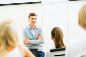 Young male teacher sat on a desk looking at the class 