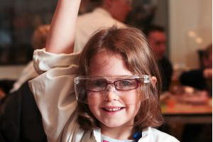 Little girl wearing safety goggles with her hand raised to ask a question 