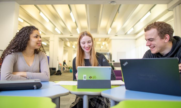 Three students are sat with their laptops talking in the Library Living Room