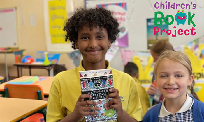 Smiling boy holding a football book in a classroom, standing next to a girl